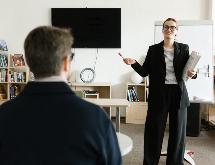 A teacher in a black blazer conducts a session with a student in a library setting.
